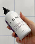Hand holding a bottle of Violet Shampoo - Alcôve Hair in a bathroom